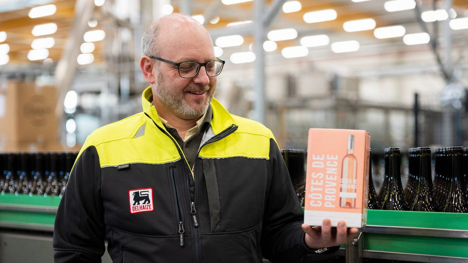 Delhaize employee holding a boxed rosé wine product beside a bottling line with glass bottles in the background.