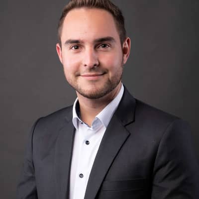 Portrait eines jungen Mannes mit kurzen, dunklen Haare. Weißes Hemd, dunkles Jacket. Er schaut in die Kamera. 