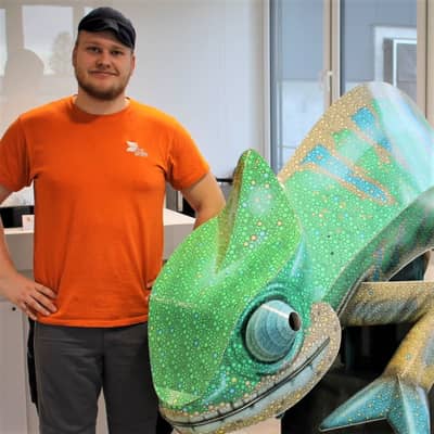 junger Mann, Mütze und orangenes T-Shirt mit Arbeitshose. Arme in die Hüfte, lächelt in die Kamera. Recht von ihm steht ein grünes Chamäleon aus Wellpappe.