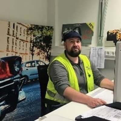 Mann mit Bart, Mütze, Gehörschutz und Sicherheitsweste am Schreibtisch. Er schaut in den PC.