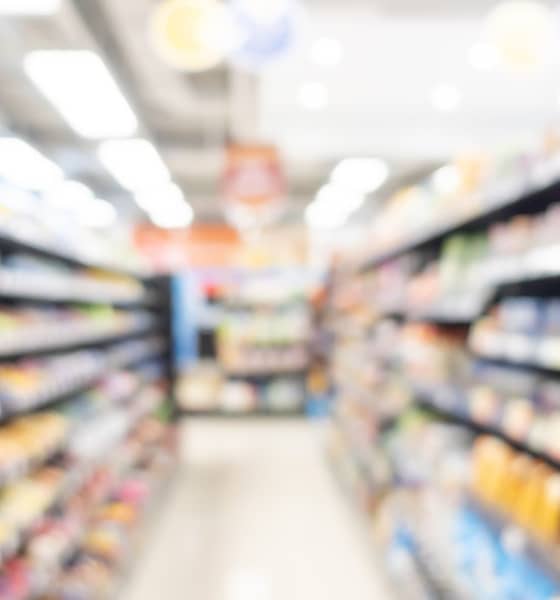 Verschwommener Gang eines Supermarkts mit vollen Regalen rechts und links sowie hinten im Bild.