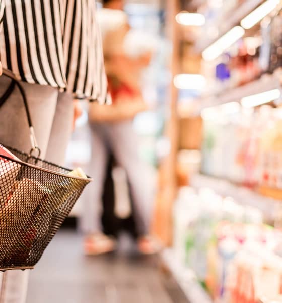 Eine Person steht vor einem Regal im Supermarkt. Im Vordergrund ist der gefüllte Einkaufskorb in der Hand zu sehen.