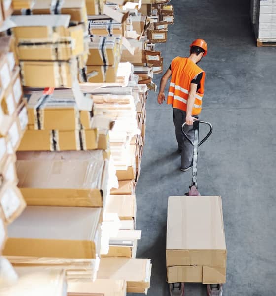 Mann im Lager voller Wellpappboxen zieht einen Hubwagen hinter sich her.