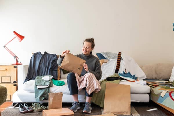 Frau sitzt auf einem weißen Sofa mit einer Papierversandtasche in der Hand. Um sie herum liegen viele Kleidungsstücke und Versandverpackungen.