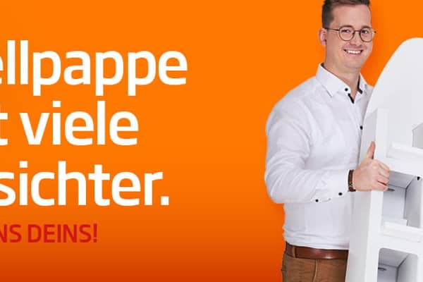 junger dunkelhaariger Mann mit weißem T-Shirt vor orangefarbenen Hintergrund lächelt in die Kamera und hält neben einem Kartonstapel einen kleinen Wellpappkarton in der Hand