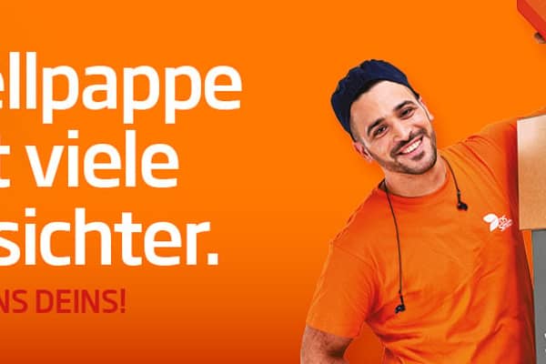  dunkelhaariger junger Mann steht links neben einem Kistenstapel aus Wellpappe, freundlich in die Kamera lächelnd, mit einem kleinen Wellpappkarton in der Hand. Mütze und Gehörschutz und oranges T-Shirt, Arbeitskleidung, vor orangefarbenem Hintergrund