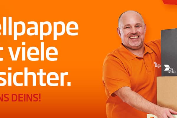 älterer weißer Mann mit kurzem Haar in orangenem T-Shirt steht links neben einem Aufsteller aus Wellpappe, freundlich in die Kamera lächelnd, mit einem kleinen Wellpappkarton in der Hand