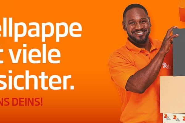 Links vom Kistenstapel aus Wellpappe steht ein junger farbiger Mann mit schwarzen Haar in orangenem T-Shirt und lächelt in die Kamera. 