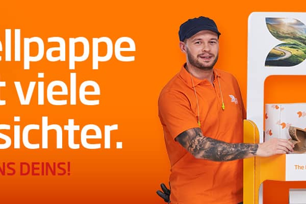 Links vom Aufsteller aus Wellpappe steht ein junger farbiger Mann mit schwarzen Haar in orangenem T-Shirt und lächelt in die Kamera. Rechts steht eine junge dunkelhaarige Frau mit weißer Bluse und dunkler Strickjacke und lächelt in die Kamera. Beide haben ein kleines Produkt aus Wellpappe in der Hand.