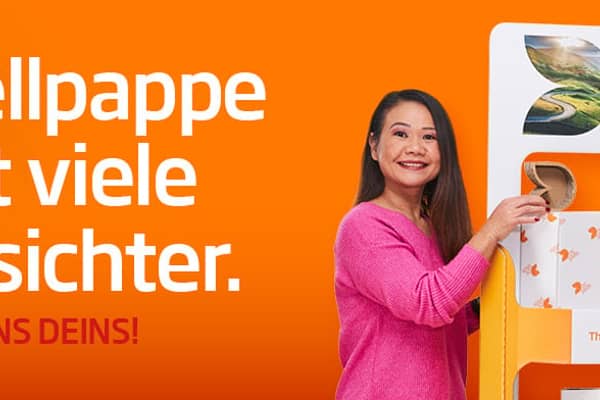  Links vom Aufsteller aus Wellpappe steht eine asiatische Frau mit langem schwarzen Haar in pinkfarbenen T-Shirt und lächelt in die Kamera. Rechts steht älterer weißer Mann mit kurzem Haar in orangenem T-Shirt steht links neben einem Kistenstapel aus Wellpappe, freundlich in die Kamera lächelnd, mit einem kleinen Wellpappkarton in der Hand