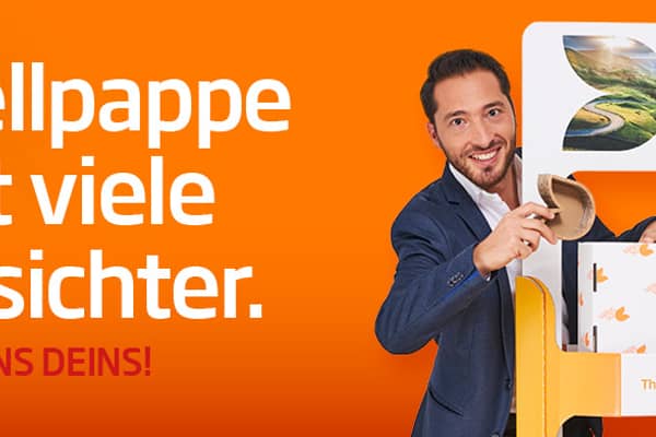 junge blonde Frau in dunklem Kleid und junger dunkelhaariger Mann mit dunklem Jacket stehen links (Frau) und rechts (Mann) von einem Wellpappaufsteller, beide halten ein Wellpappkarton in der Hand und lächeln in die Kamera.