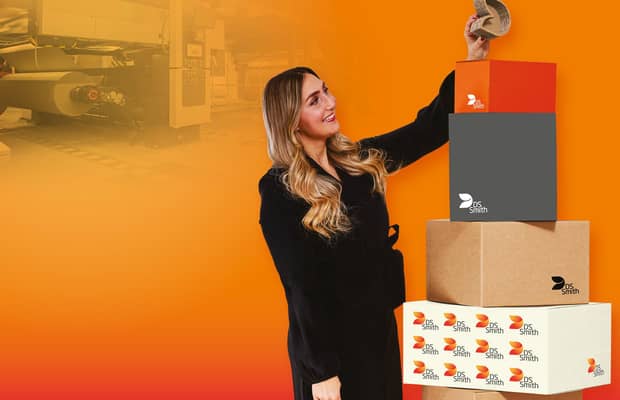 junge blonde Frau in dunklem Kleid steht links von einem Wellpappaufsteller und hält einen kleinen Wellpappkarton in der Hand und lächelt.