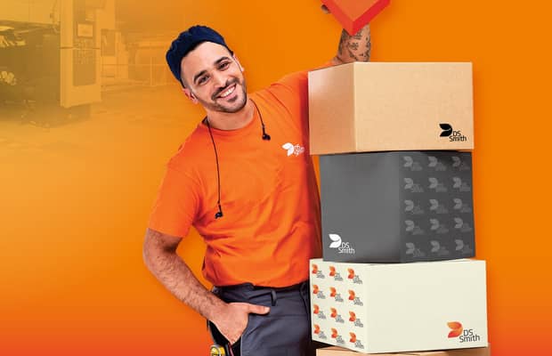 dunkelhaariger junger Mann links neben einem Kistenstapel aus Wellpappe, freundlich in die Kamera lächelnd, mit einem kleinen Wellpappkarton in der Hand. Mütze und Gehörschutz und oranges T-Shirt, Arbeitskleidung, vor orangefarbenem Hintergrund