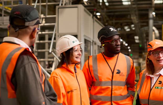 A diverse group of packaging plant employees are standing in a semicircle and looking at someone outside of the shot. They are all wearing safety equipment branded International Paper and DS Smith.