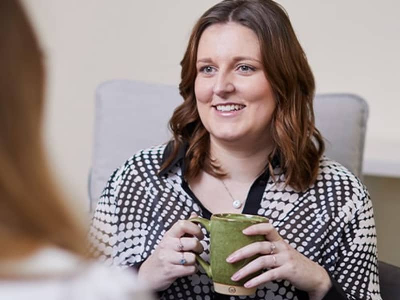 A woman holding a cup and talking to a female colleague.