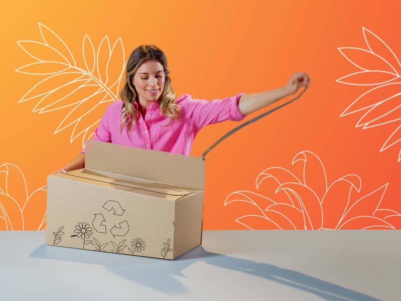 Eine blonde Frau öffnet eine braune Verpackung welche auf einem grauen Tisch steht. Die Verpackung ist mit Blumen und einem Recyclingsymbol bedruckt.
