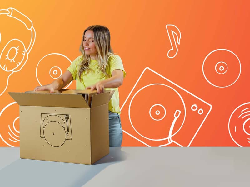 Eine blonde Frau hantiert mit einer auf einem Tisch stehenden Verpackung. Auf der braunen Verpackung ist ein Schallplattenspieler zu sehen und der Hintergrund ist orange mit verschiedenen Musikzeichnungen.