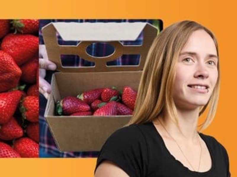 Frau im Vordergrund schaut nach vorne, während im Hintergrund Wellpappschalen mit Erdbeeren präsentiert sind.