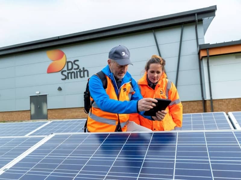 Eine Frau und ein Mann schauen zusammen auf ein Tablet. Beide haben Warnjacken an und stehen zwischen Fotovoltaikanlagen vor einem Gebäude mit DS Smith Logo.