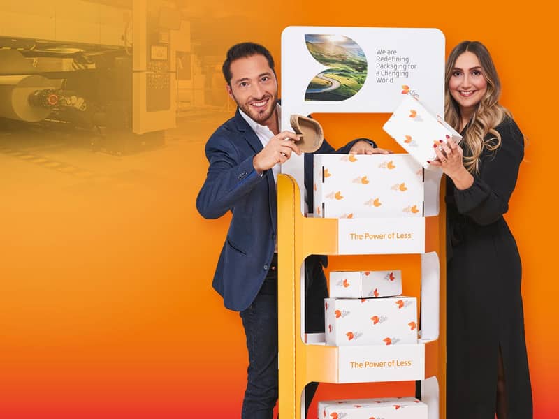 junge blonde Frau in dunklem Kleid und junger dunkelhaariger Mann mit dunklem Jacket stehen links (Mann) und rechts (Frau) von einem Wellpappaufsteller, beide halten ein Wellpappkarton in der Hand und lächeln in die Kamera.