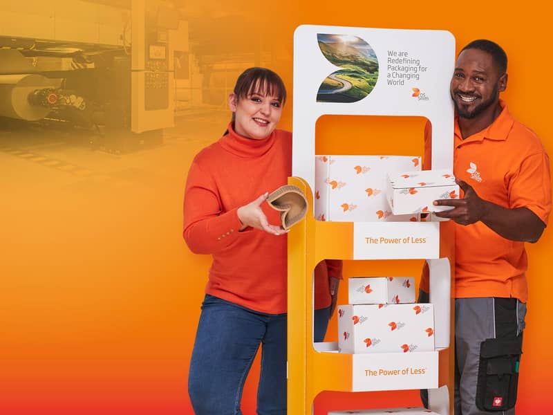  Rechts vom Aufsteller aus Wellpappe steht ein junger farbiger Mann mit schwarzen Haar in orangenem T-Shirt und lächelt in die Kamera. Links steht eine junge dunkelhaarige Frau mit orangenem Pullover und lächelt in die Kamera. Beide haben ein kleines Produkt aus Wellpappe in der Hand.