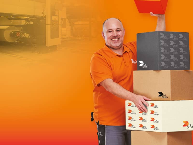  älterer weißer Mann mit kurzem Haar in orangenem T-Shirt steht links neben einem Kistenstapel aus Wellpappe, freundlich in die Kamera lächelnd, mit einem kleinen Wellpappkarton in der Hand