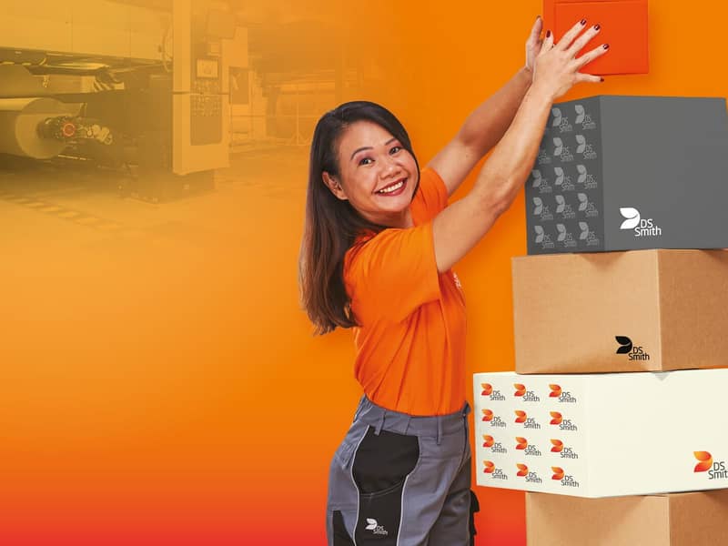 asiatische Frau mit langen Haaren steht links eines Kartonstapels aus Wellpappe vor orangefarbenem Hintergrund und lächelt in die Kamera. Sie trägt ein orangefarbenes T-Shirt was deutlich Arbeitskleidung der Produktion ist. Sie hält mit beiden Händen einen kleinen Karton aus Wellpappe und stellt diesen auf den Stapel oben drauf.