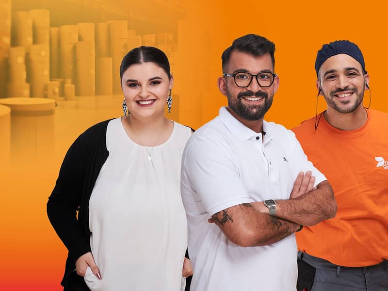 Eine Gruppe von 3 Mitarbeitenden vor orangenem Hintergrund. In der Mitte ein junger Mann mit weißem T-Shirt und vor der Brust verschränkten Armen. Es handelt sich um legere Bekleidung für das Büro (Design). Links von ihm eine junge dunkelhaarige Frau mit Zopf in weißer Bluse mit dunkler Strickjacke im Büro Look. Rechts von ihm ein dunkelhaariger junger Mann. Mütze und Gehörschutz und oranges T-Shirt, Arbeitskleidung. Links von der Gruppe steht der Employer Branding Claim „Wellpappe hat viele Gesichter. Zeig uns Deins.“