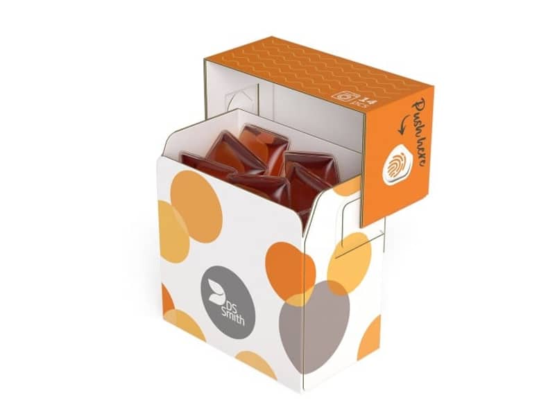 Geöffnete Verpackung für Waschmittel mit weißem Unterteil und gelben, orangen und grauen Kreisen und orangenem Deckel. In der Packung befinden sich rote Waschmittel-Pods.