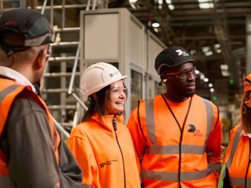 A diverse group of packaging plant employees are standing in a semicircle and looking at someone outside of the shot. They are all wearing safety equipment branded International Paper and DS Smith.