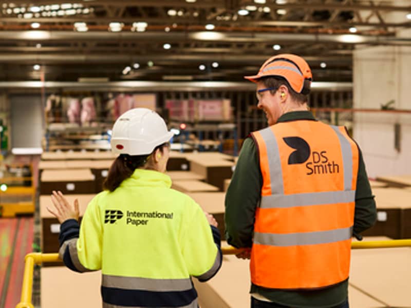 Two packaging plant workers, a man and a woman, seen from behind as they look over the plant. They are wearing reflective safety vests branded International Paper and DS Smith.