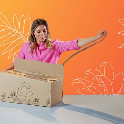 Eine blonde Frau öffnet eine braune Verpackung welche auf einem grauen Tisch steht. Die Verpackung ist mit Blumen und einem Recyclingsymbol bedruckt.
