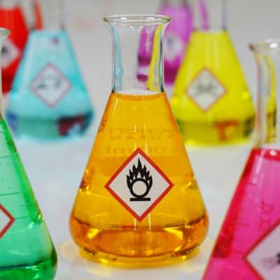 Mehrere Laborgläser gefüllt mit Gefahrgutetiketten und bunten Flüssigkeiten.