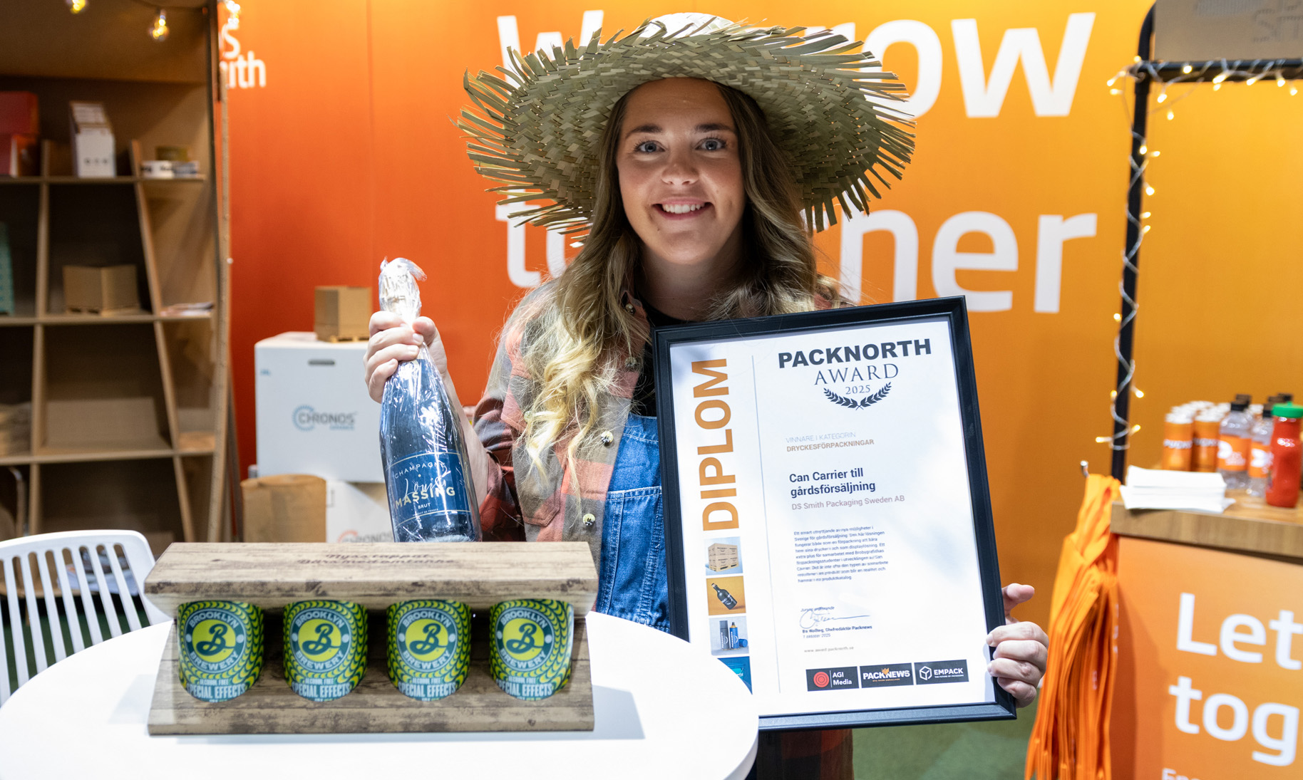 Emelie Karlsson från DS Smith håller upp diplom och champagne efter att ha tagit emot Packnorth Award 2025 för Can Carrier i kategorin dryckesförpackningar. Framför henne står den prisbelönta förpackningen med fyra burkar.