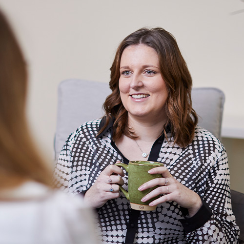A woman holding a cup and talking to a female colleague.