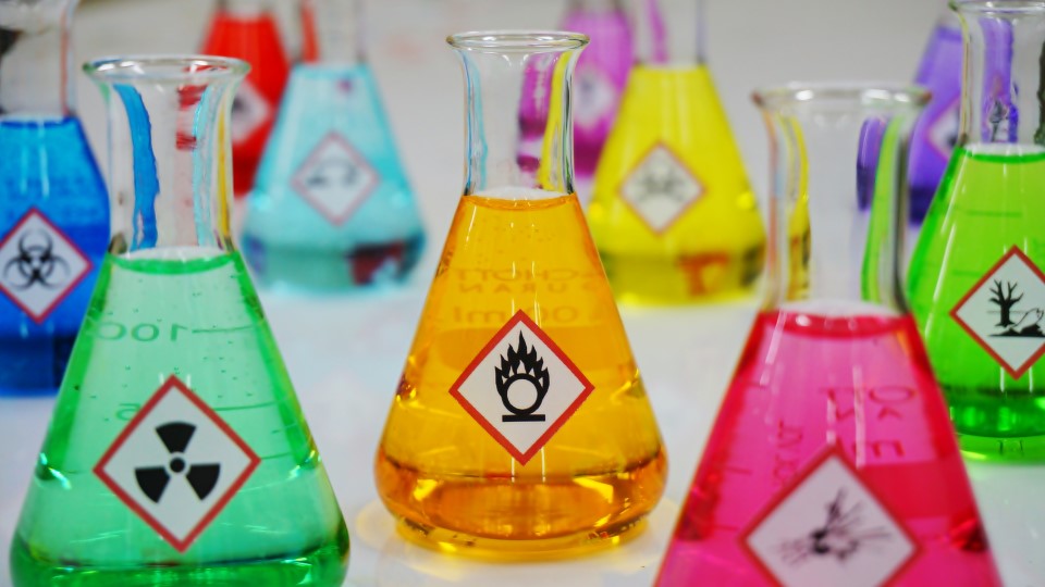 Mehrere Laborgläser mit Warnetiketten und gefüllt mit bunten Flüssigkeiten.