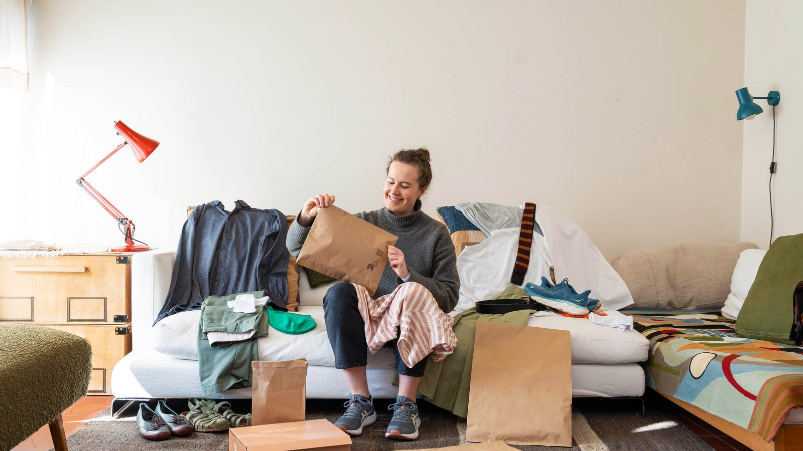 Frau sitzt auf einem weißen Sofa mit einer Papierversandtasche in der Hand. Um sie herum liegen viele Kleidungsstücke und Versandverpackungen.