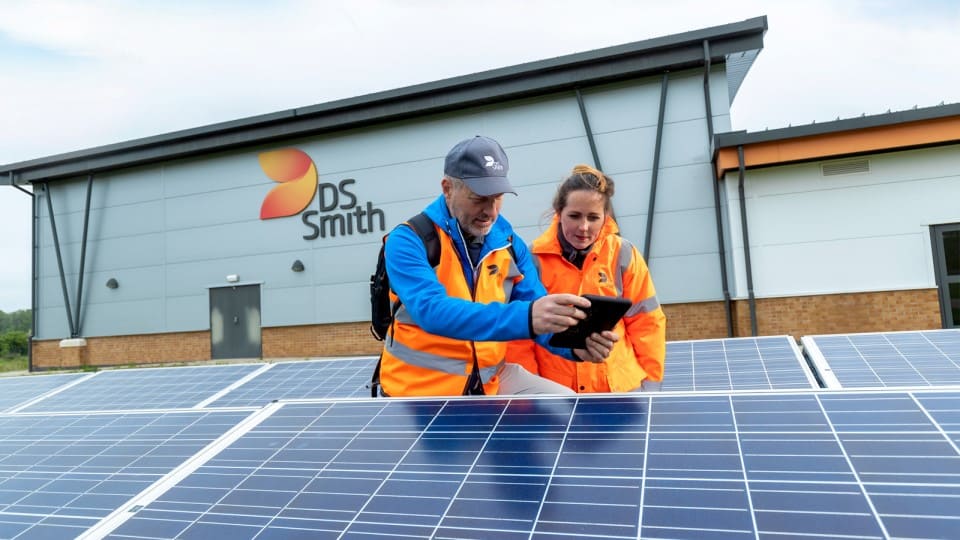 Eine Frau und ein Mann schauen zusammen auf ein Tablet. Beide haben Warnjacken an und stehen zwischen Fotovoltaikanlagen vor einem Gebäude mit DS Smith Logo.