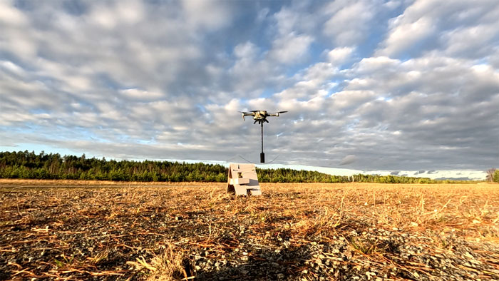 drone kuljettaa aaltopahvista jalustaa ja raivauspanoslaatikkoa pellolla