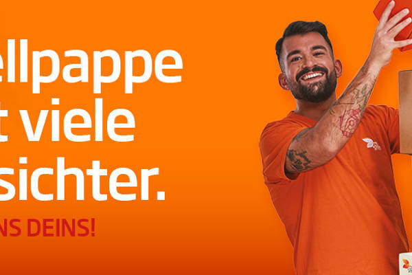 asiatische Frau mit langen Haaren steht seitlich eines Kartonstapels aus Wellpappe vor orangefarbenem Hintergrund und lächelt in die Kamera. Sie trägt ein orangefarbenes T-Shirt was deutlich Arbeitskleidung der Produktion ist.