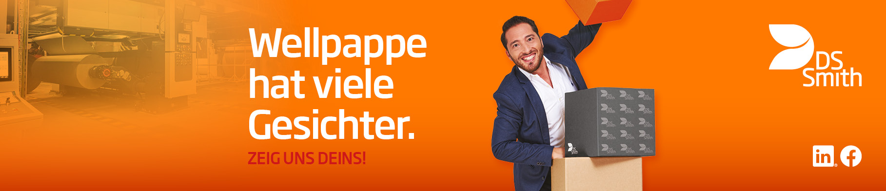 junger dunkelhaariger Mann mit dunklem Jacket steht links hinter einem Wellpappaufsteller und hält ein Wellpappkarton in der Hand und lächelt in die Kamera.