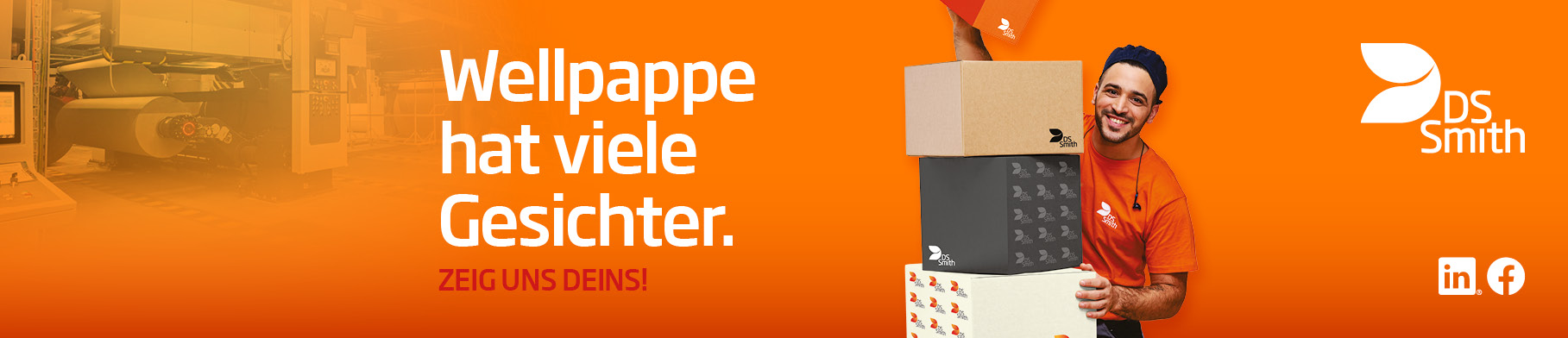 dunkelhaariger junger Mann steht rechts neben und halb hinter einem Kistenstapel aus Wellpappe, freundlich in die Kamera lächelnd, mit einem kleinen Wellpappkarton in der Hand. Mütze und Gehörschutz und oranges T-Shirt, Arbeitskleidung, vor orangefarbenem Hintergrund