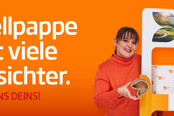 junges Mädchen mit dunklen langen Haaren zum Zopf steht links von einem Aufsteller aus Wellpappe, freundlich in die Kamera lächelnd. Rechts vom Aufsteller steht ein junger farbiger Mann mit schwarzen Haar in organgenem TShirt und lächelt in die Kamera. Beide stehen vor einem orangefarbenen Hintergrund