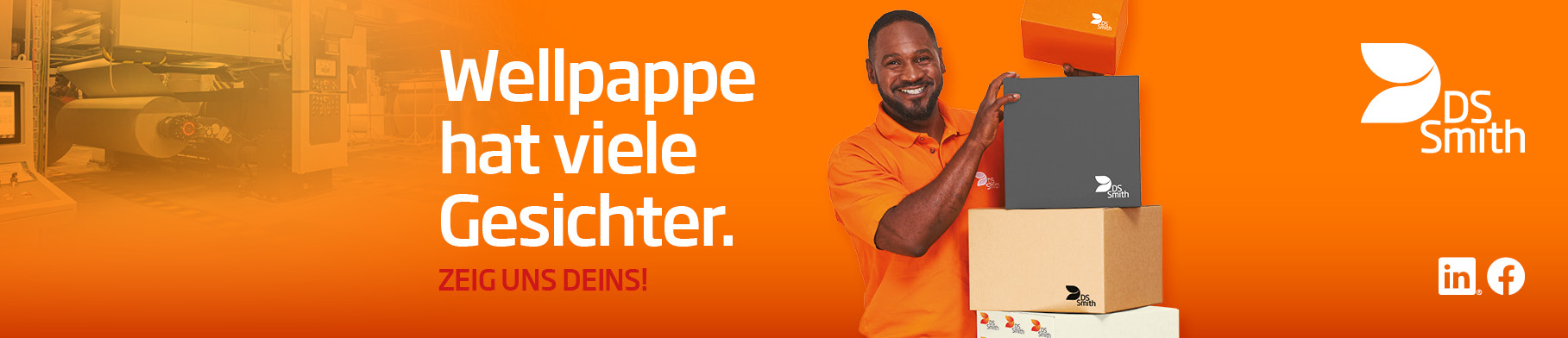 Links vom Kistenstapel aus Wellpappe steht ein junger farbiger Mann mit schwarzen Haar in orangenem T-Shirt und lächelt in die Kamera. 