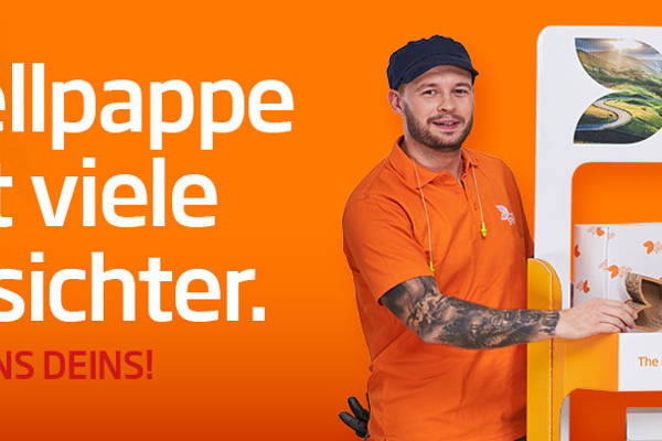 Links vom Aufsteller aus Wellpappe steht ein junger farbiger Mann mit schwarzen Haar in orangenem T-Shirt und lächelt in die Kamera. Rechts steht eine junge dunkelhaarige Frau mit weißer Bluse und dunkler Strickjacke und lächelt in die Kamera. Beide haben ein kleines Produkt aus Wellpappe in der Hand.