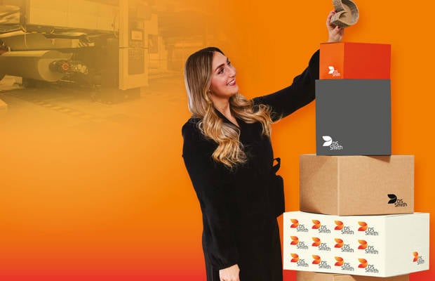 junge blonde Frau in dunklem Kleid steht links von einem Wellpappaufsteller und hält einen kleinen Wellpappkarton in der Hand und lächelt.