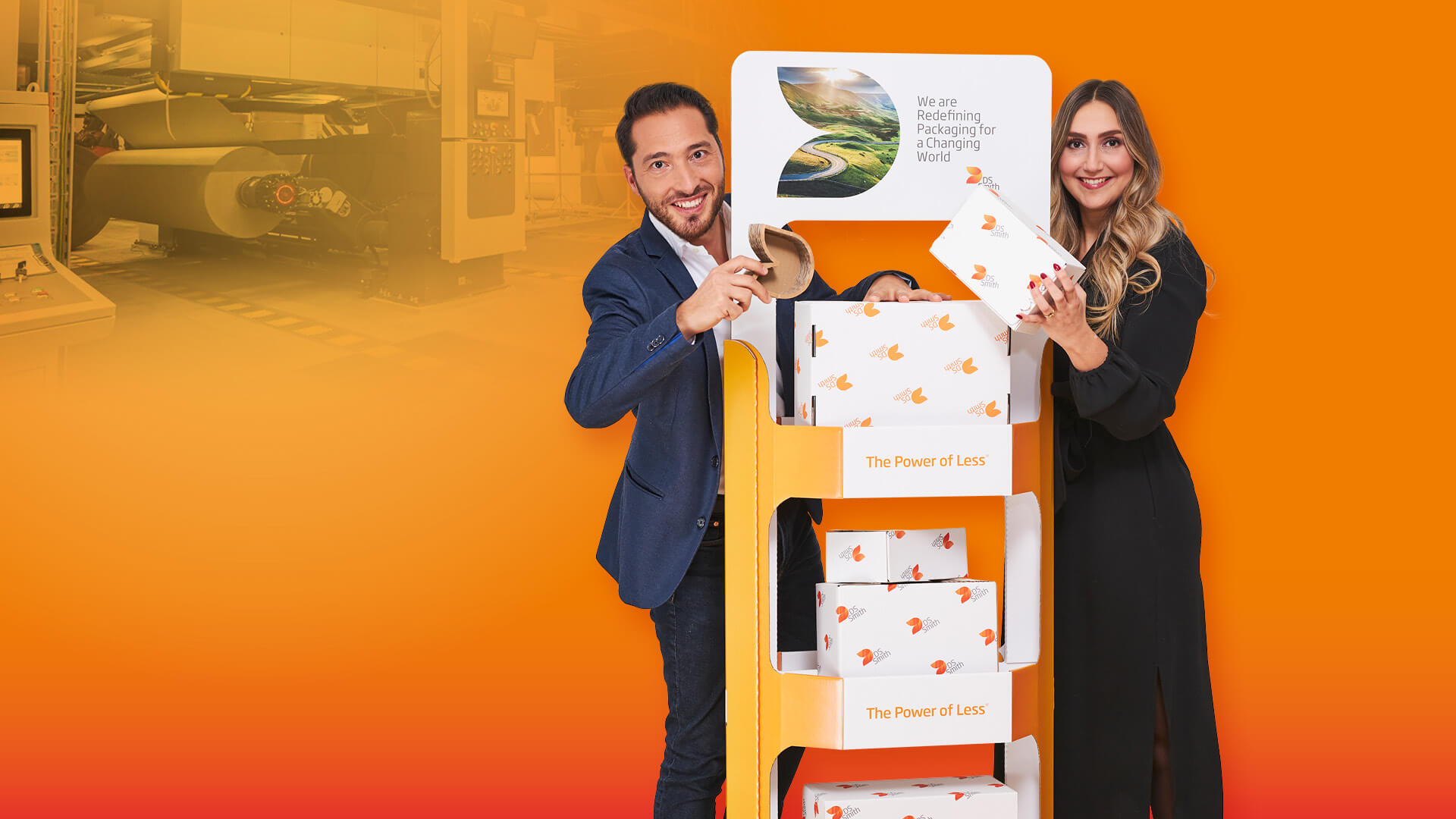 junge blonde Frau in dunklem Kleid und junger dunkelhaariger Mann mit dunklem Jacket stehen links (Mann) und rechts (Frau) von einem Wellpappaufsteller, beide halten ein Wellpappkarton in der Hand und lächeln in die Kamera.