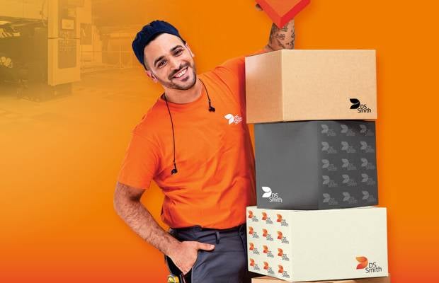 dunkelhaariger junger Mann links neben einem Kistenstapel aus Wellpappe, freundlich in die Kamera lächelnd, mit einem kleinen Wellpappkarton in der Hand. Mütze und Gehörschutz und oranges T-Shirt, Arbeitskleidung, vor orangefarbenem Hintergrund