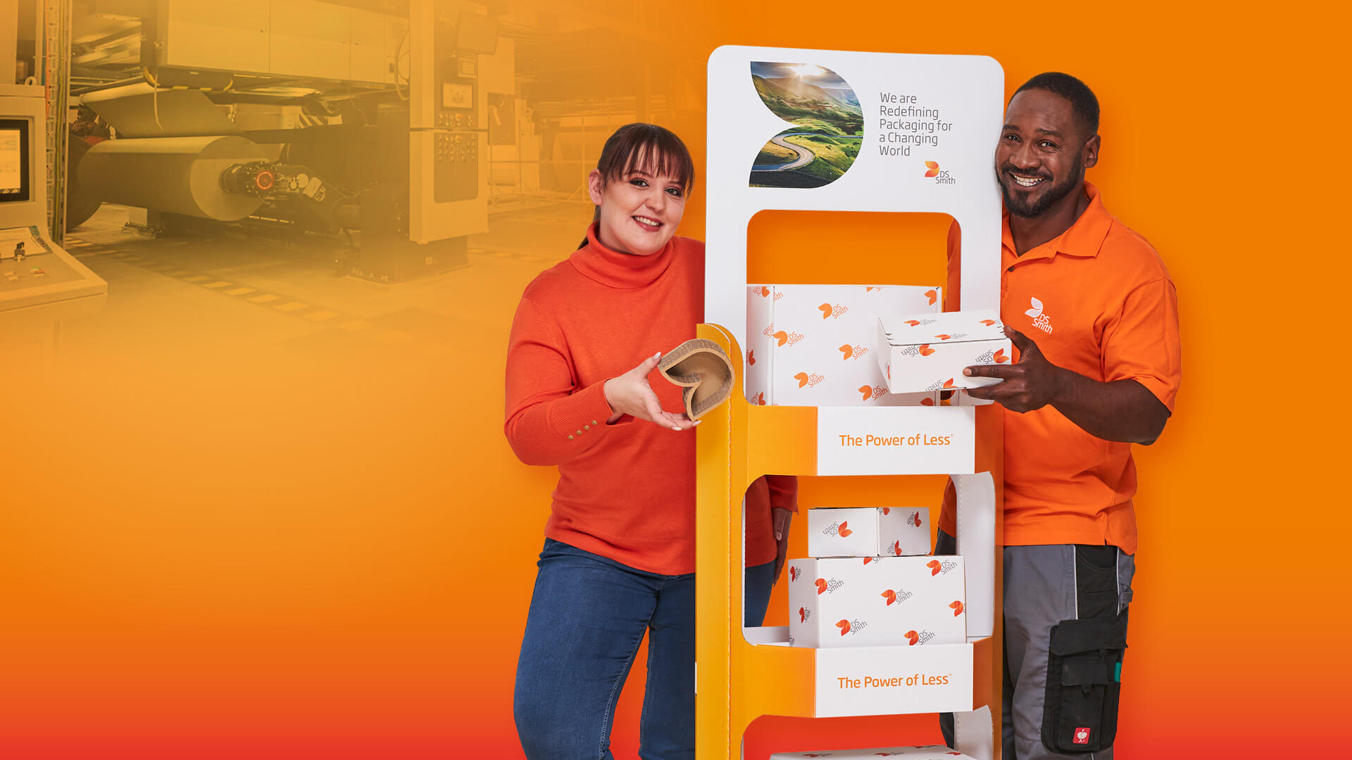  Rechts vom Aufsteller aus Wellpappe steht ein junger farbiger Mann mit schwarzen Haar in orangenem T-Shirt und lächelt in die Kamera. Links steht eine junge dunkelhaarige Frau mit orangenem Pullover und lächelt in die Kamera. Beide haben ein kleines Produkt aus Wellpappe in der Hand.