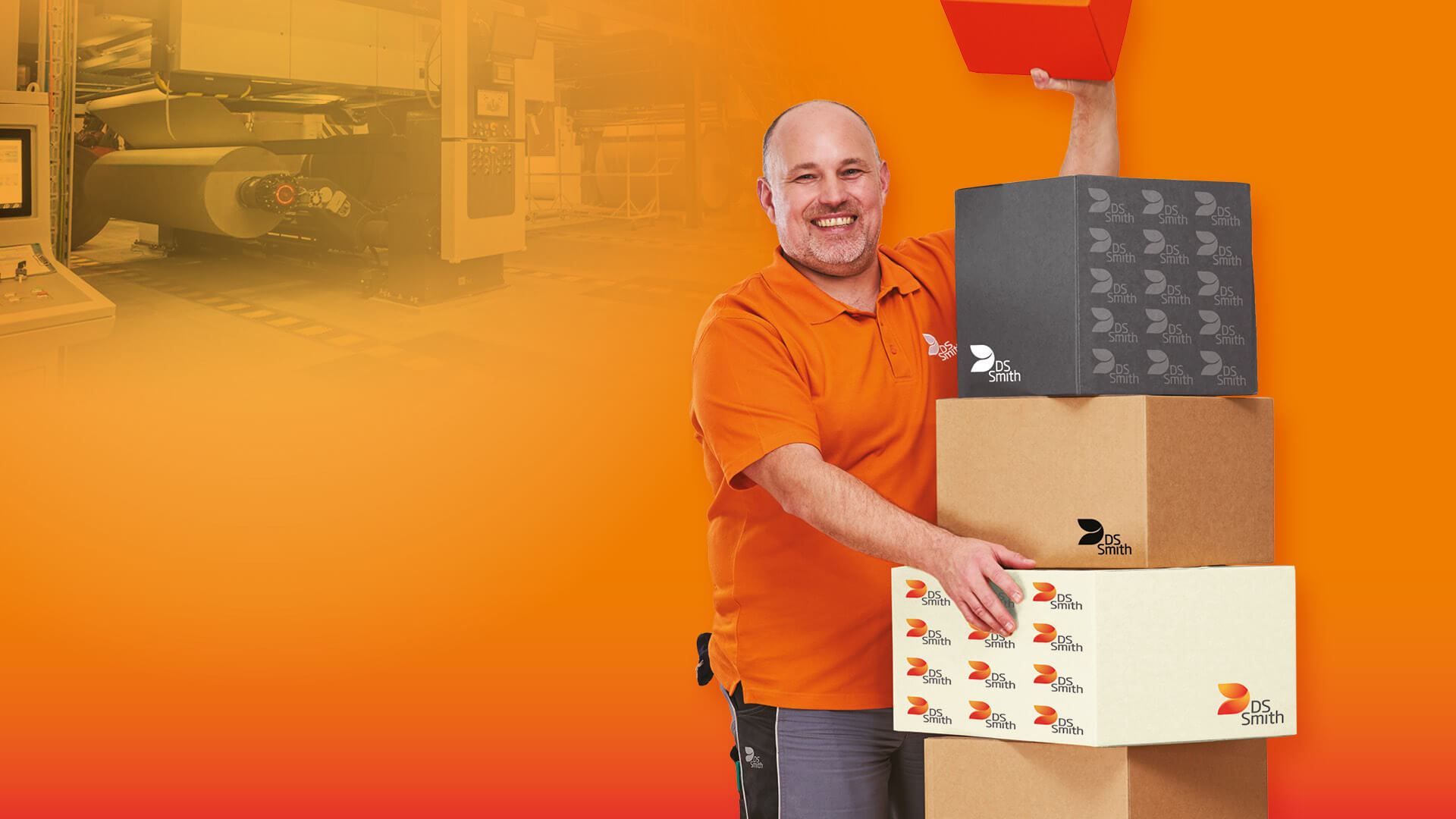  älterer weißer Mann mit kurzem Haar in orangenem T-Shirt steht links neben einem Kistenstapel aus Wellpappe, freundlich in die Kamera lächelnd, mit einem kleinen Wellpappkarton in der Hand